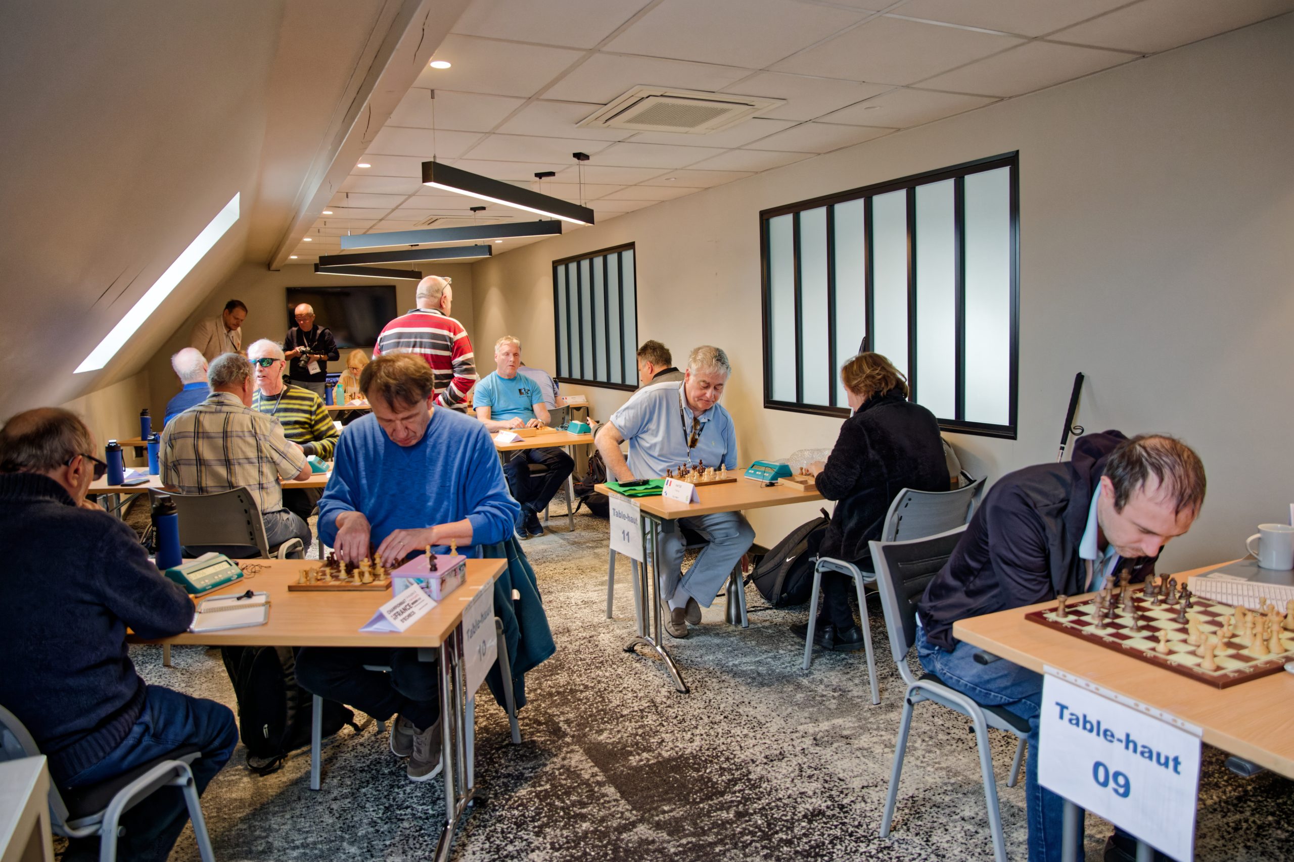 Salle de jeu du haut remplie d'un peu plus de 10 personnes en train de jouer aux échecs.