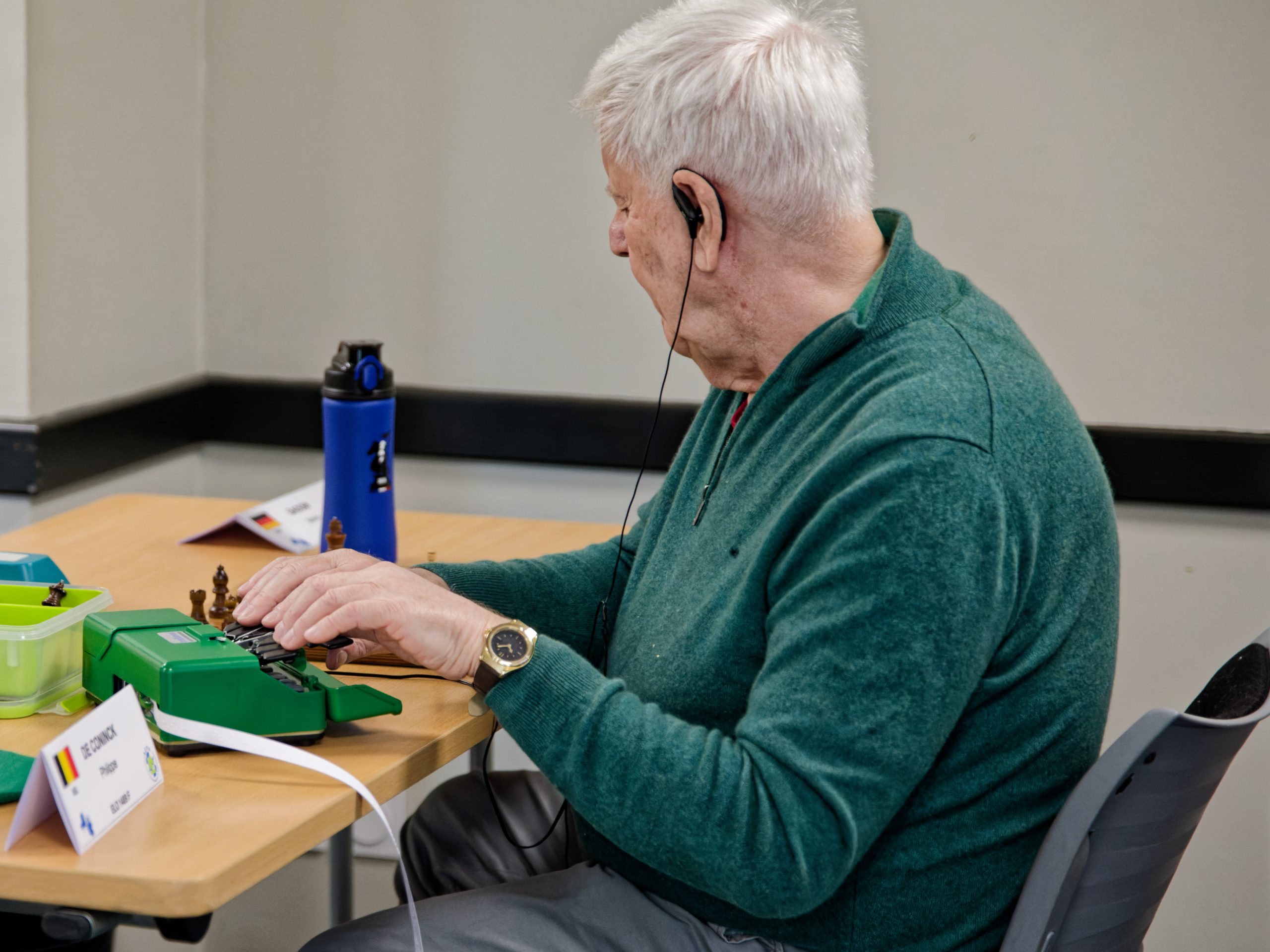 Martin GADOW, oreillette fixée à son oreille, note un coup sur une sorte de machine à écrire Braille. A l'arrière de la machine, une bandelette de papier en sort et tombe jusqu'au sol.