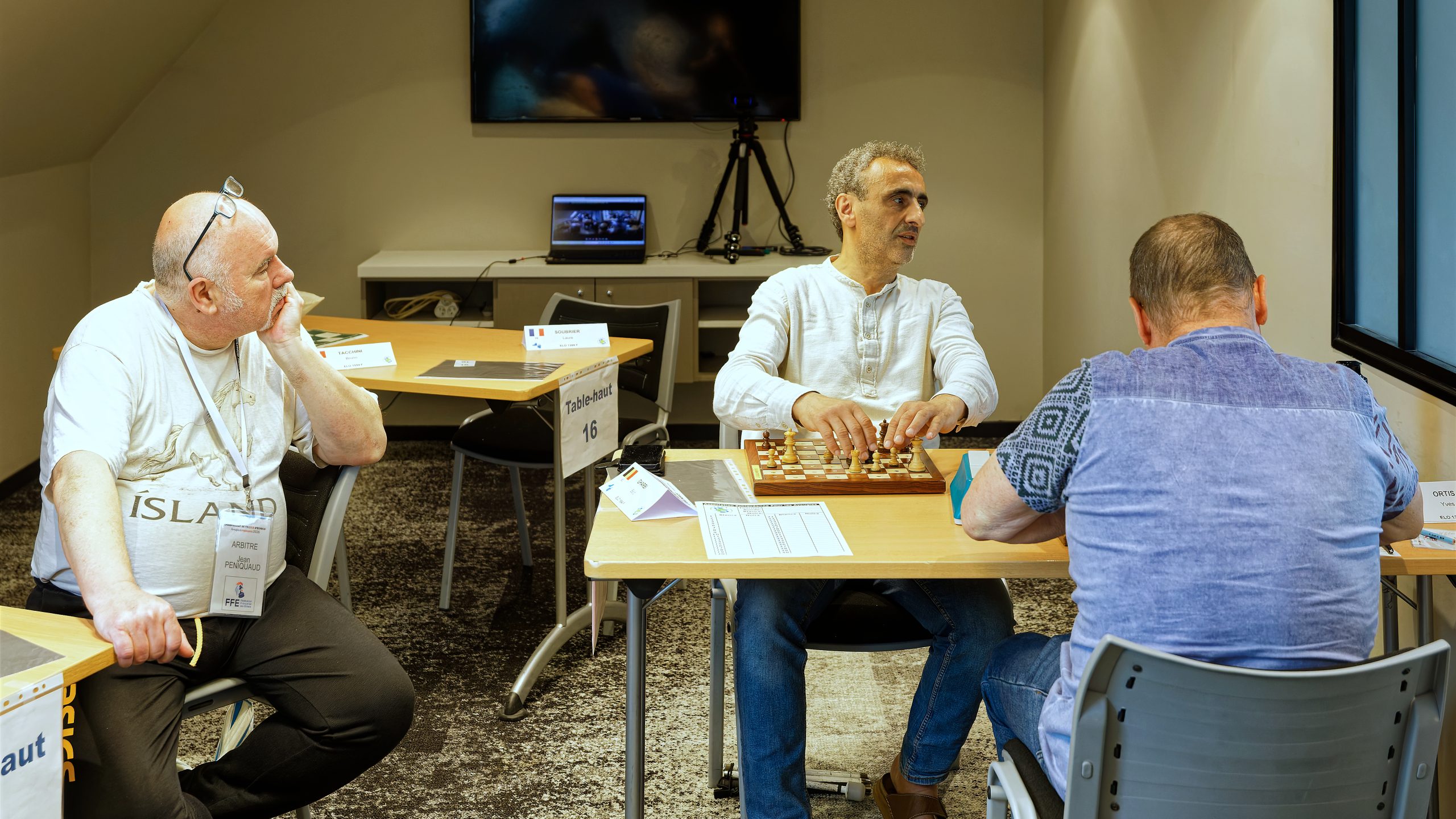 Jean, l'arbitre, surveille une partie. Le temps semble lui paraître long, il est assis sur une chaise d'une table terminée à côté. Il ne reste plus grand monde dans la salle.