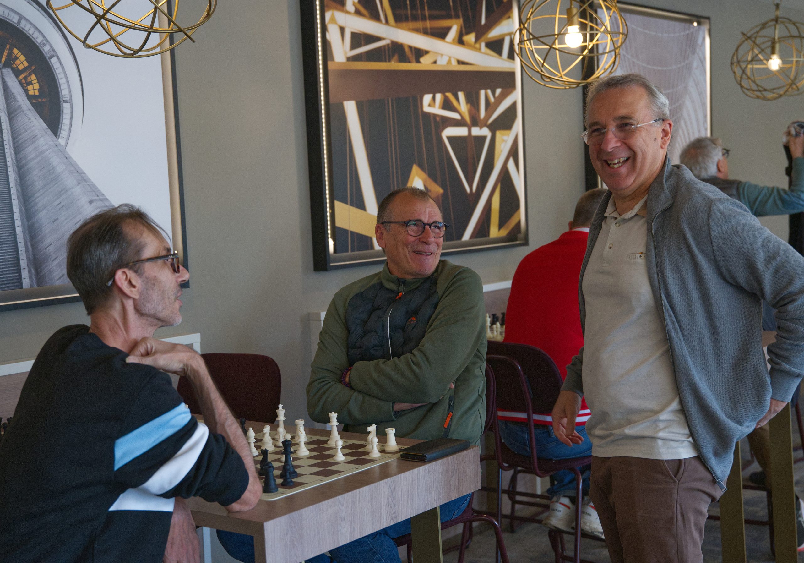 Dans la salle de restauration, Gérard joue une partie contre Miguel, bénévole pendant ce tournoi. L'ambiance semble détendue et Stéphane rigole avec eux à côté de l'échiquier.