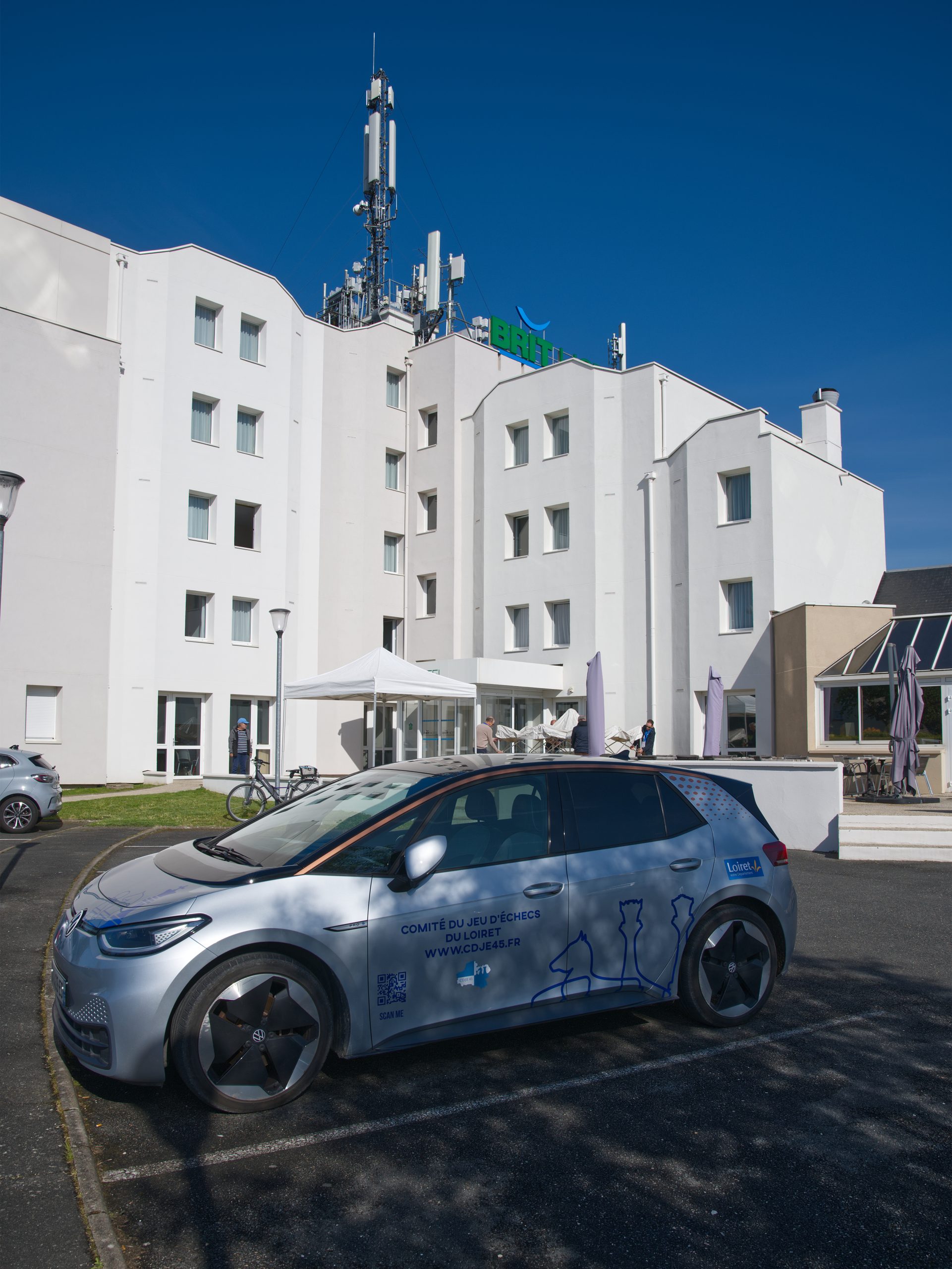 Une voiture électrique avec un flocage sur le côté indiquant "Comité départemental du jeu d'échecs du Loiret" des silhouettes de pièces finissent d'habiller la carosserie. La voiture est stationnée sur le parking de l'hôtel que l'on aperçoit en arrière plan.