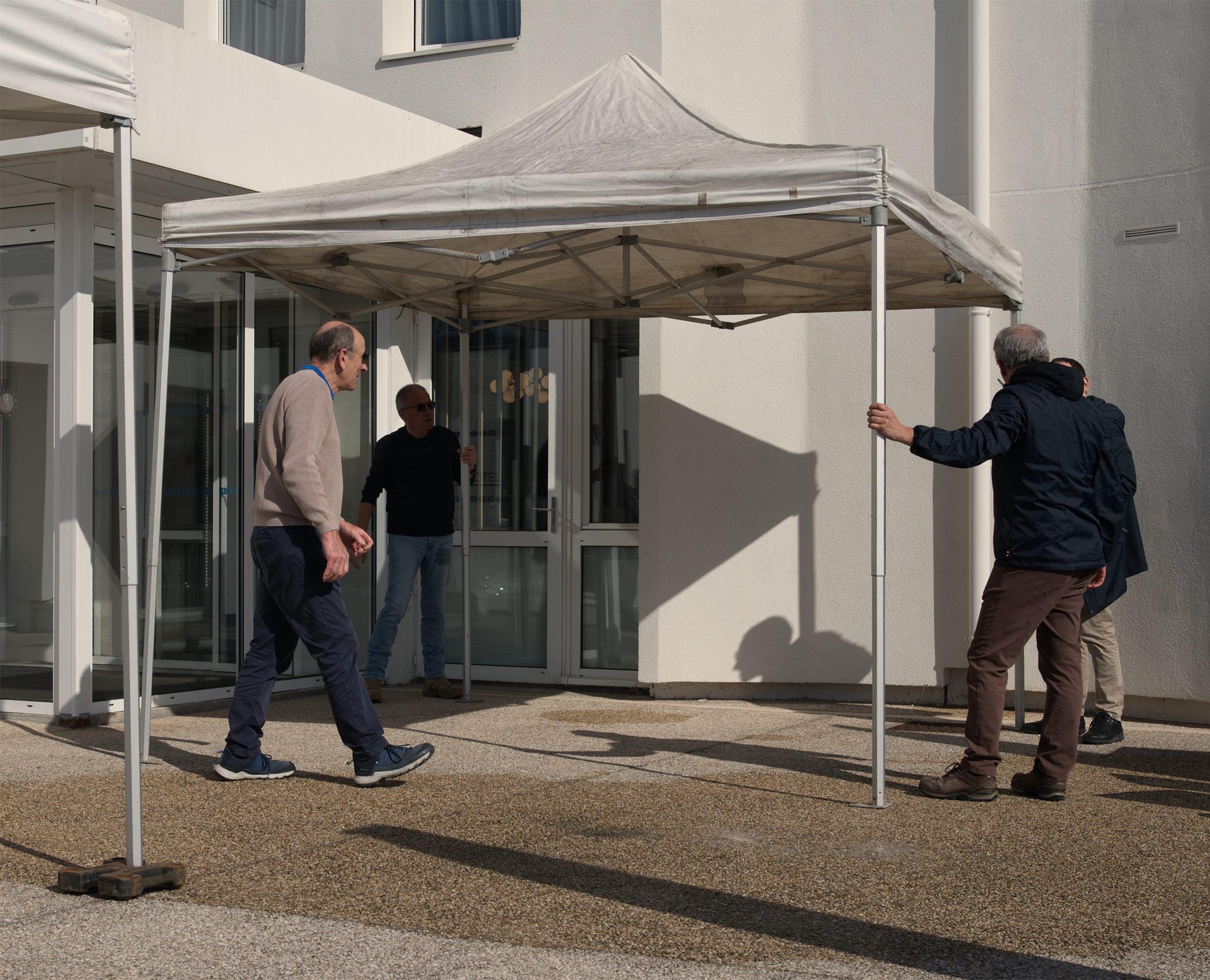 4 personnes déploient un abri pour installer les stands prévus pour l'après-midi. Le sol est encore mouillé, la météo est incertaine.