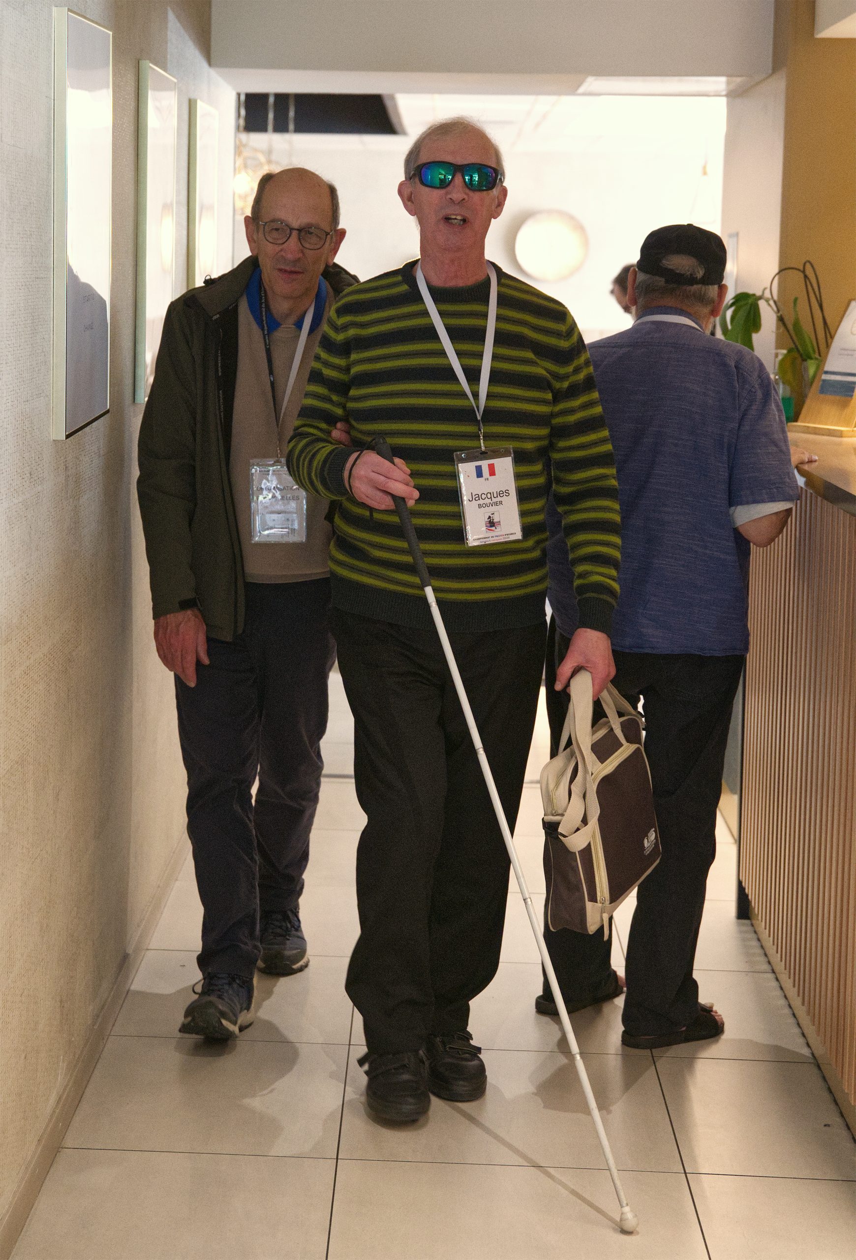 Jacques BOUVIER marche dans le couloir le long de l'accueil, suivi par Eric VIGNELLES. A droite, André CLAUZEL attend au bar.