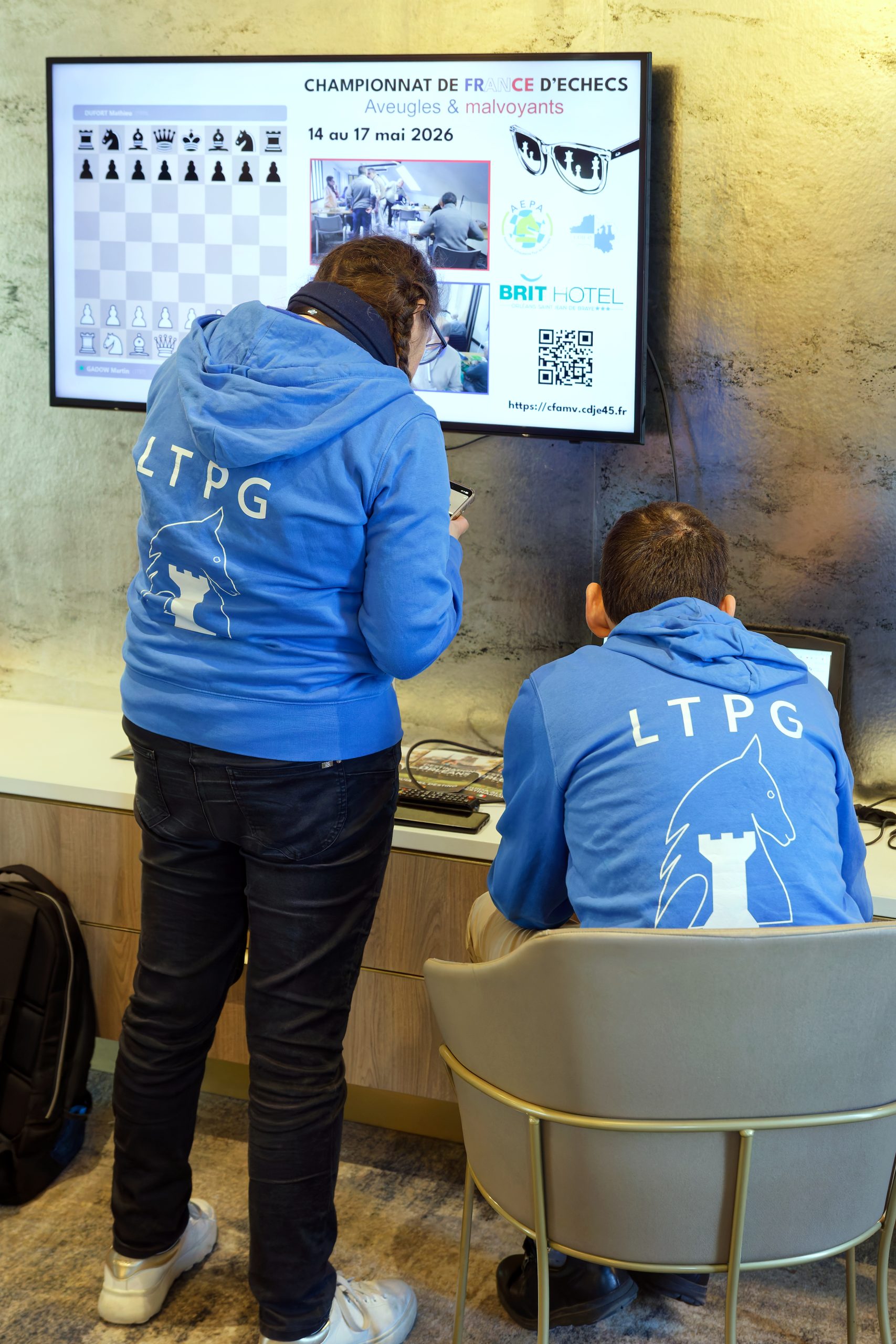 Jour 2, Aurélie et Jérémy, arborant fièrement les sweat-shirt bleus du club LTPG, lancent la retransmission sur l'écran du salon. Suite à l'échec de retransmission de la veille, une application a été développée pendant la nuit pour pouvoir retransmettre également les coups d'un échiquier, suivi avec son téléphone par Sébastien, le bénévole qui s'est occupé de la retransmission.
